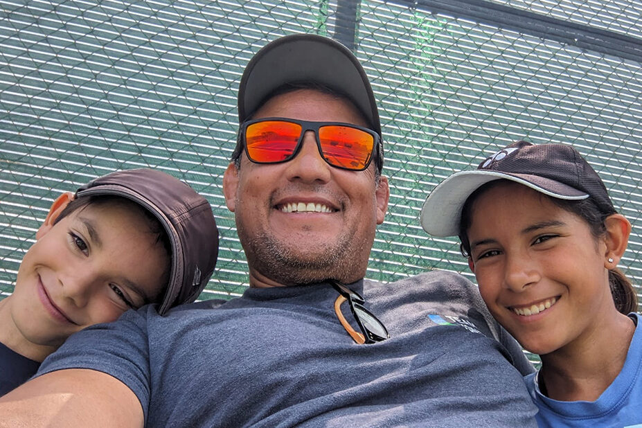 Kiana and Paolo Huerta smiling with their father and tennis coach Rafael Huerta on the tennis court.