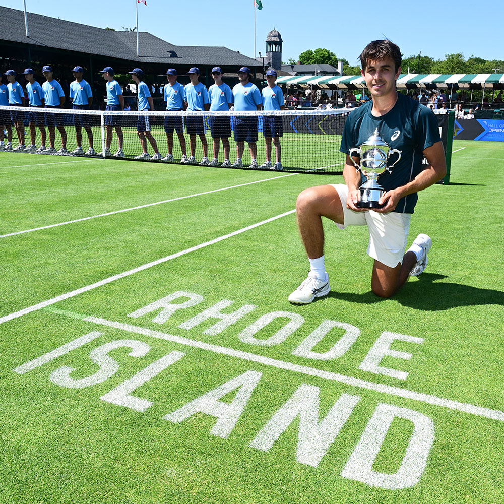 Zach Svajda kneels on the grass court holding his ATP Challenger 125 trophy with "Rhode Island" painted on the court at the Hall of Fame Open in Newport.