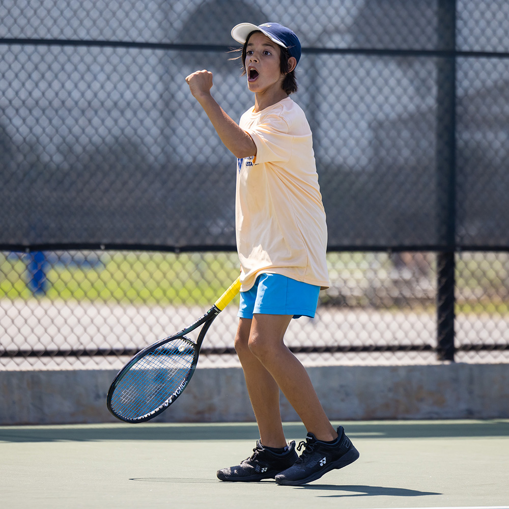 Paolo Huerta celebrates with a fist pump during a tennis match at the 2024 Kats Kup in Southern California. Wearing a light yellow shirt and holding his racquet, Huerta captures the intensity and energy of junior tennis competition.