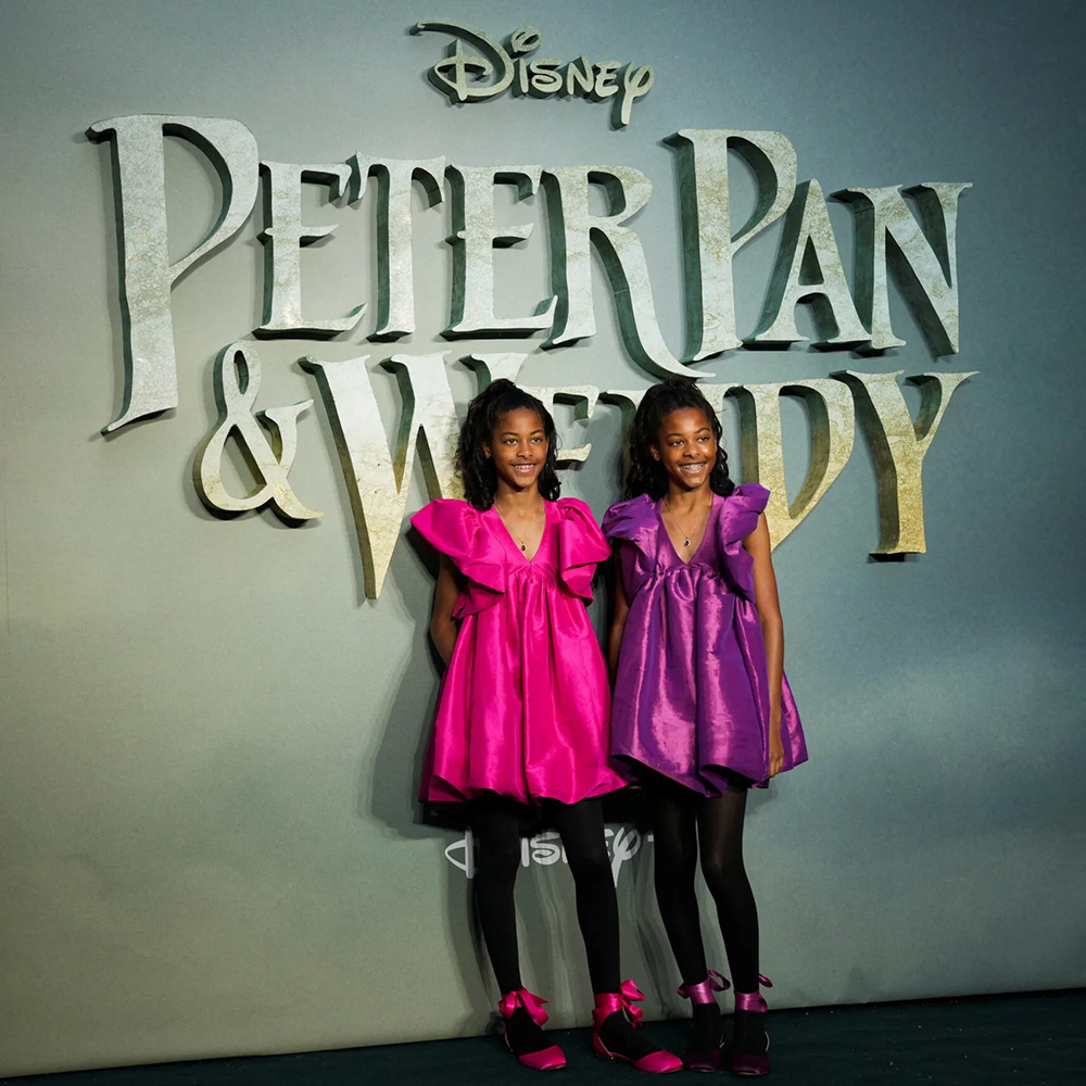 Skyler and Kelsey Yates, twin sisters, stand in front of a Peter Pan & Wendy backdrop wearing matching pink and purple dresses at the film’s world premiere in London.