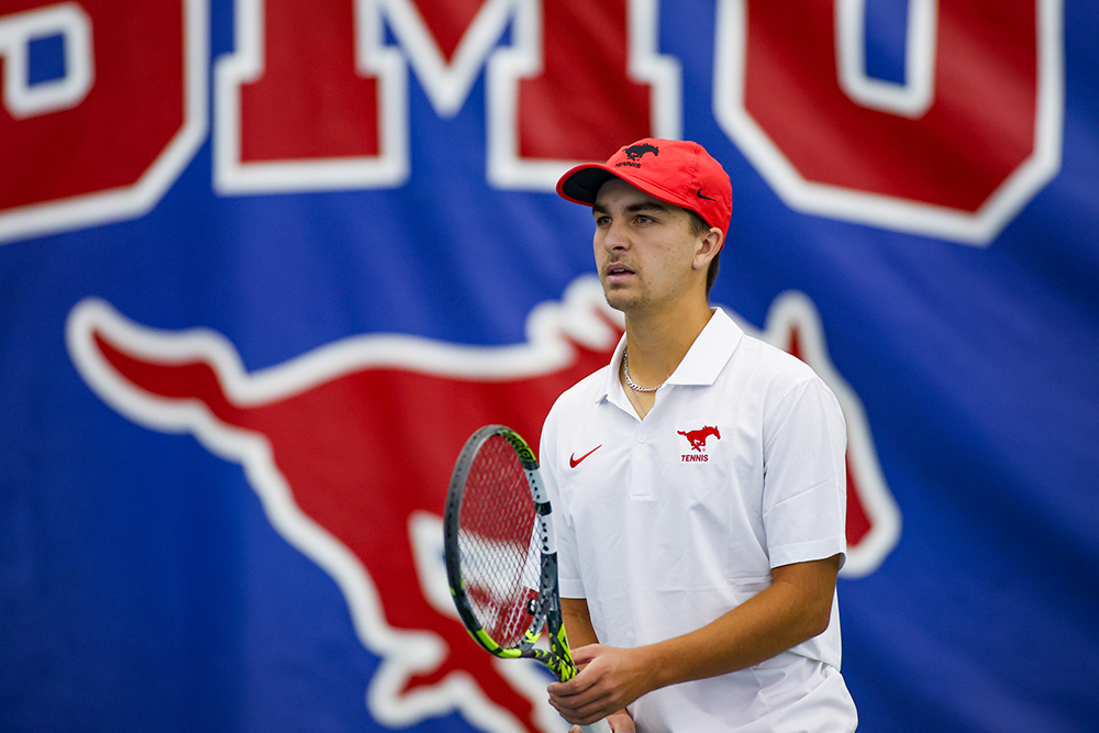 Tennis player Trevor Svajda stands on a court holding his racket, looking focused during a match. He is wearing a white SMU Tennis polo shirt and a red cap featuring the SMU Mustang logo. A large blue banner with the red and white SMU logo and mustang mascot is visible in the background.