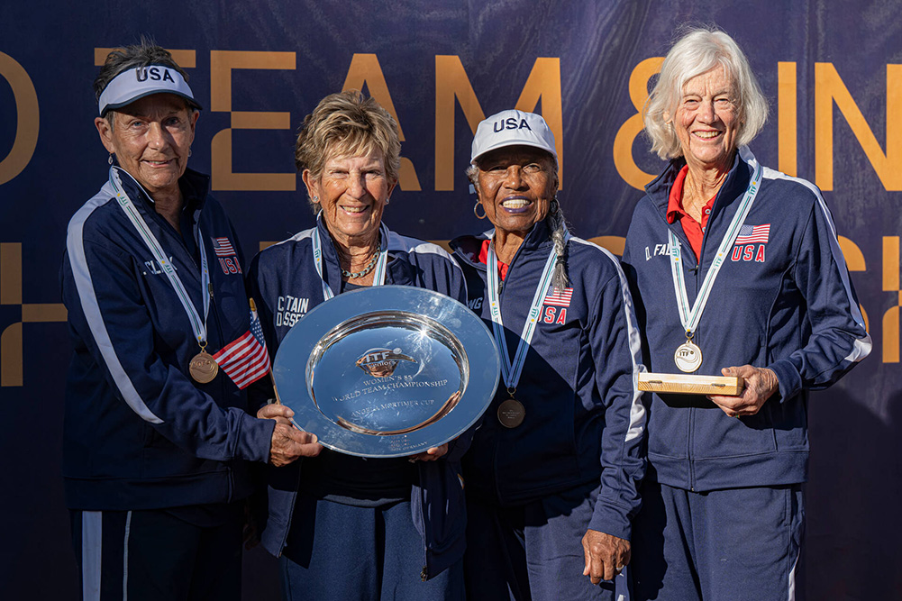 The Team USA Women’s 85 champions pose together with their trophy. From left to right: Susanne Clark, Dorothy Wasser, Roz King, and Donna Fales. They are wearing navy blue Team USA tracksuits and gold medals. Dorothy and Roz hold a large silver trophy plate inscribed with "Angela Mortimer Cup."
