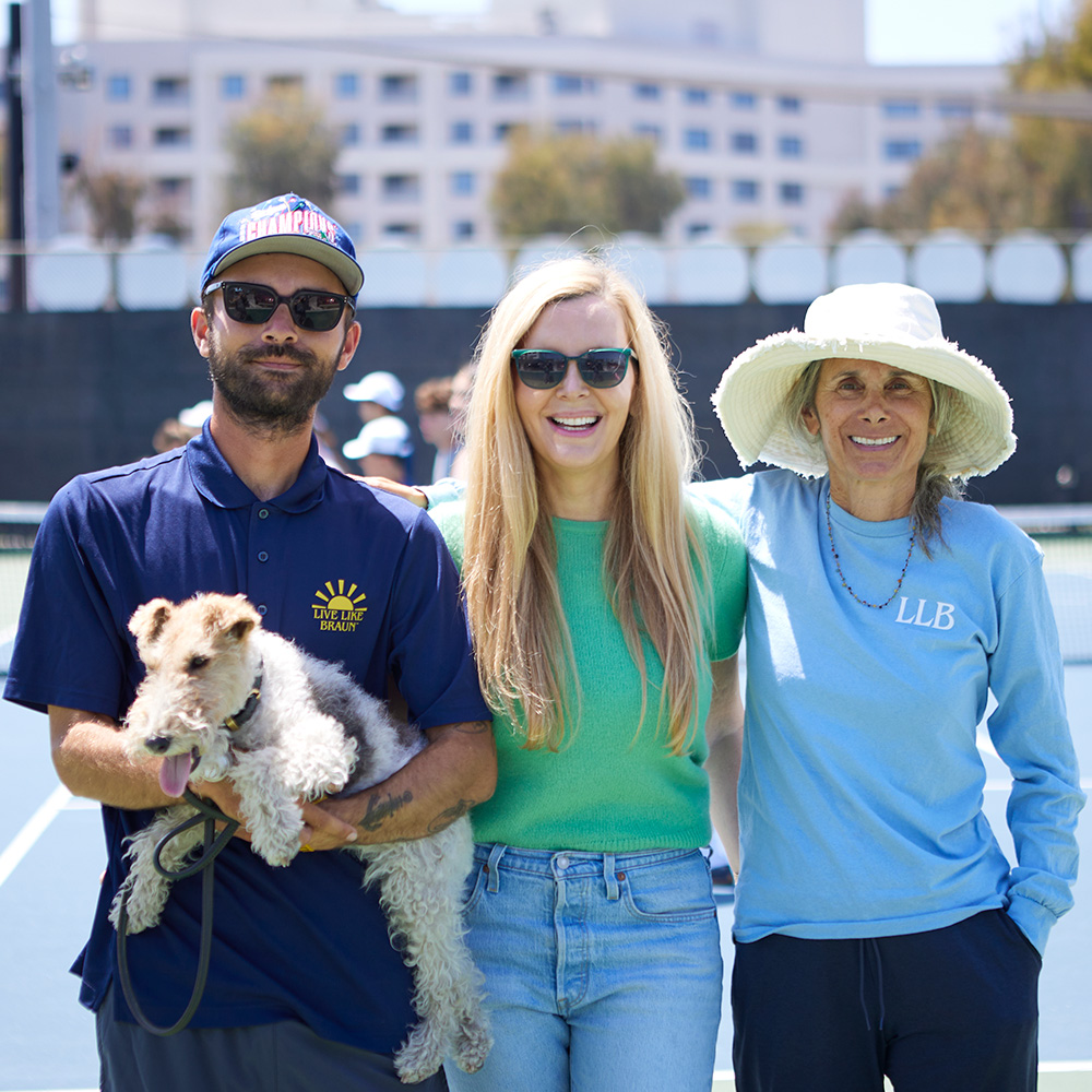 Henry Mahdessian, Susan Zinn, and Debbie Mahdessian smile together on a sunny tennis court. Henry holds their dog, Daisy, a wire-haired terrier, while Susan wears a green shirt and sunglasses, and Debbie wears a light blue long-sleeved shirt and a white sun hat.