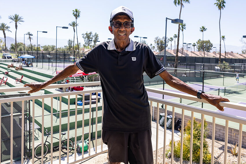 A portrait of Gordon ZawTun smiling while standing on an outdoor balcony overlooking a tennis facility in Palm Desert. He is wearing a black Wilson polo shirt, dark shorts, a white and black baseball cap, and glasses. Behind him, several green and purple tennis courts are visible under a bright, clear sky, accented by tall palm trees and distant mountains.
