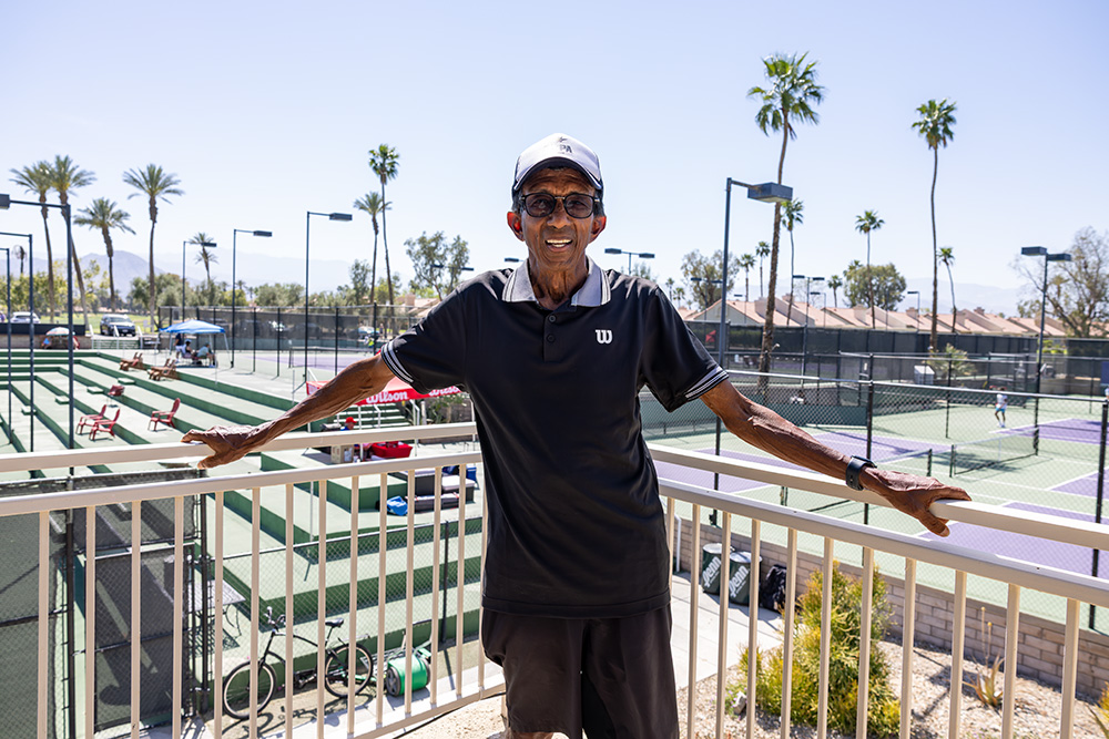 A portrait of Gordon ZawTun smiling while standing on an outdoor balcony overlooking a tennis facility in Palm Desert. He is wearing a black Wilson polo shirt, dark shorts, a white and black baseball cap, and glasses. Behind him, several green and purple tennis courts are visible under a bright, clear sky, accented by tall palm trees and distant mountains.