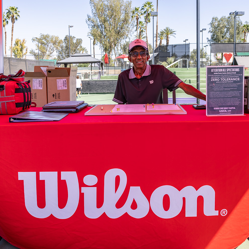 Tennis tournament director Gordon ZawTun smiles while sitting behind a registration desk draped in a bright red Wilson tablecloth. He is wearing a dark polo shirt and a white baseball cap. On the table in front of him are tournament papers, a red gear bag, and a "Zero Tolerance" spectator policy sign. In the sunny background, green tennis courts and palm trees are visible under a clear sky.