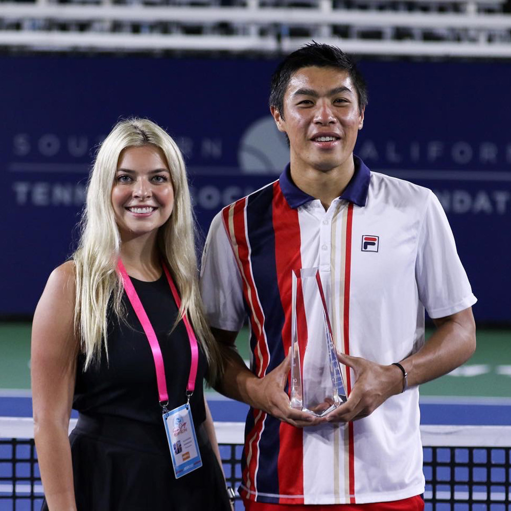 Photographer Lexie Wanninger and tennis player Brandon Nakashima stand side-by-side on an outdoor hard court after Brandon won the San Diego Open. Lexie, on the left, smiles brightly while wearing a black sleeveless top, a black skirt, and a pink tournament credential around her neck. Brandon, on the right, wears a white, red, and navy striped Fila polo shirt and holds a clear crystal trophy. In the background, a dark blue stadium wall displays the text "Southern California Tennis Association Foundation."