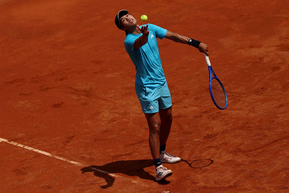 Learner Tien is captured in the middle of a service motion on a red clay court at the Geneva Open. He tosses a yellow tennis ball into the air with his left hand while holding a blue racket in his right. He is wearing a turquoise Adidas kit consisting of a t-shirt and matching shorts, along with a black visor and a black wristband. A sharp shadow is cast behind him on the bright, sunlit clay.