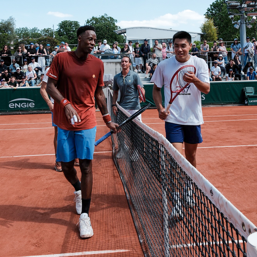Tennis stars Gael Monfils and Brandon Nakashima walk alongside the net during a practice session at Roland Garros in May 2026. Monfils, on the left, is wearing a burnt orange t-shirt, blue shorts, and white tennis shoes, smiling as he talks with Nakashima. Brandon, on the right, wears a white t-shirt and navy shorts while holding his red racket. The scene is set on the iconic red clay courts in Paris, with a crowd of spectators watching from the stands in the background. This tournament marks a poignant milestone as Monfils' 19th and final appearance at his home Grand Slam before his retirement at the end of the 2026 season.