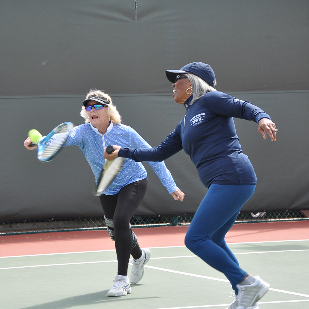 Roz King and Donna Fales are captured in a dynamic action shot during a doubles match. Donna (left) wearing a blue patterned long-sleeve shirt and sunglasses, prepares to hit a forehand, while Roz (right) in a navy long-sleeve top and cap, moves quickly across the court in support.