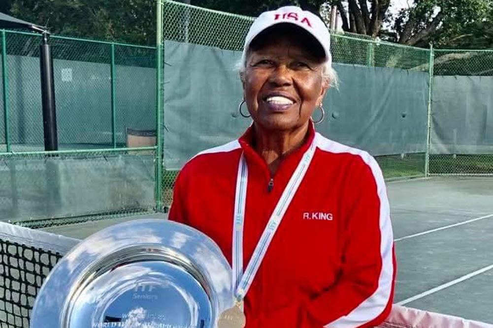 A portrait of Roz King standing on a tennis court, proudly holding a large silver championship trophy plate. She is wearing a red and white athletic jacket, a white "USA" cap, and her gold winner's medal.