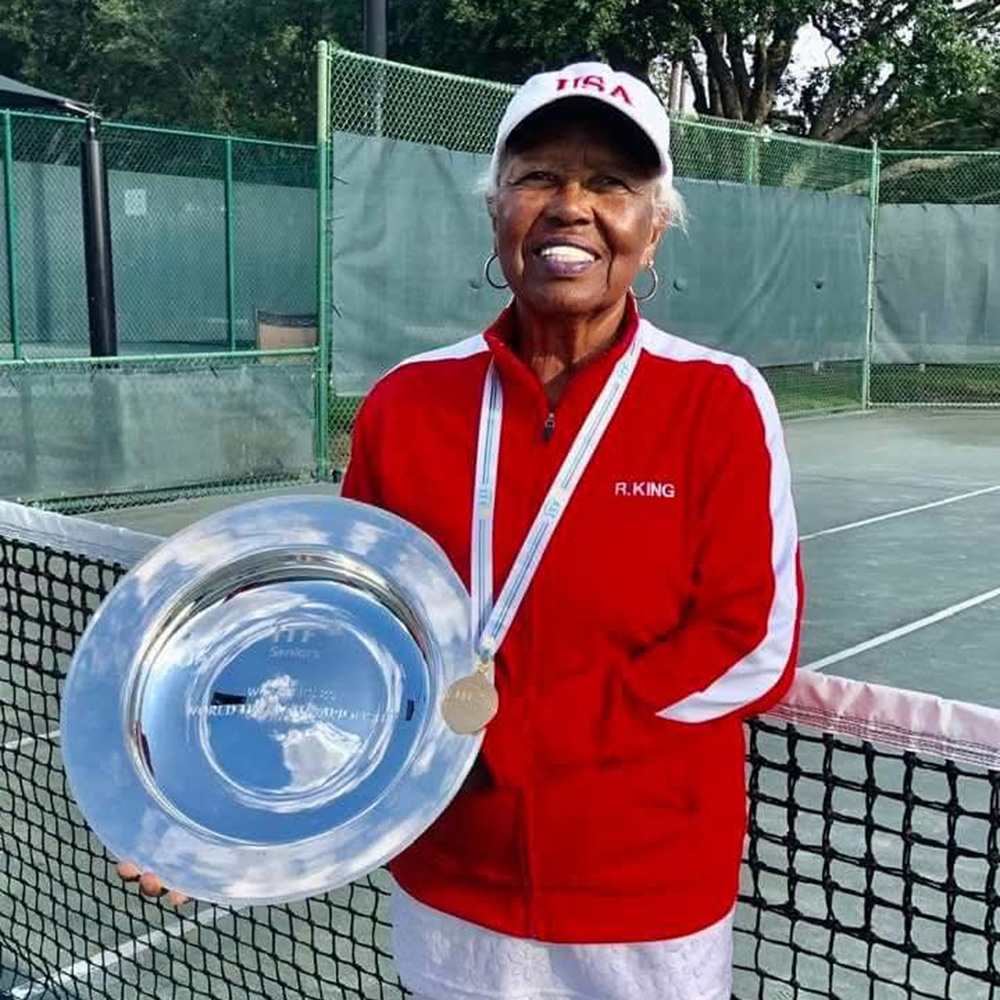 A portrait of Roz King standing on a tennis court, proudly holding a large silver championship trophy plate. She is wearing a red and white athletic jacket, a white "USA" cap, and her gold winner's medal.