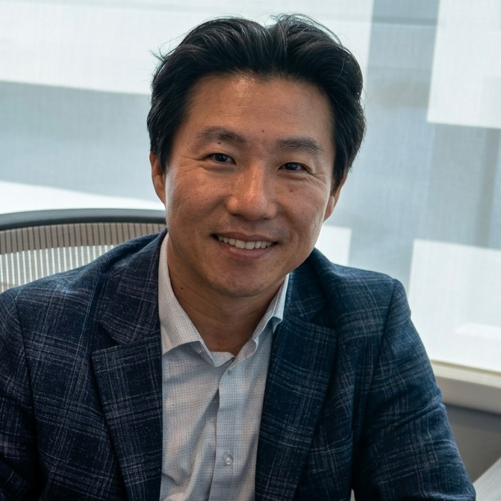 A professional headshot of Dr. KyoungBin Im smiling while seated in an office chair. He is wearing a dark blue plaid blazer over a light-colored button-down shirt. The background is bright and airy with modern office windows, providing soft, natural light.