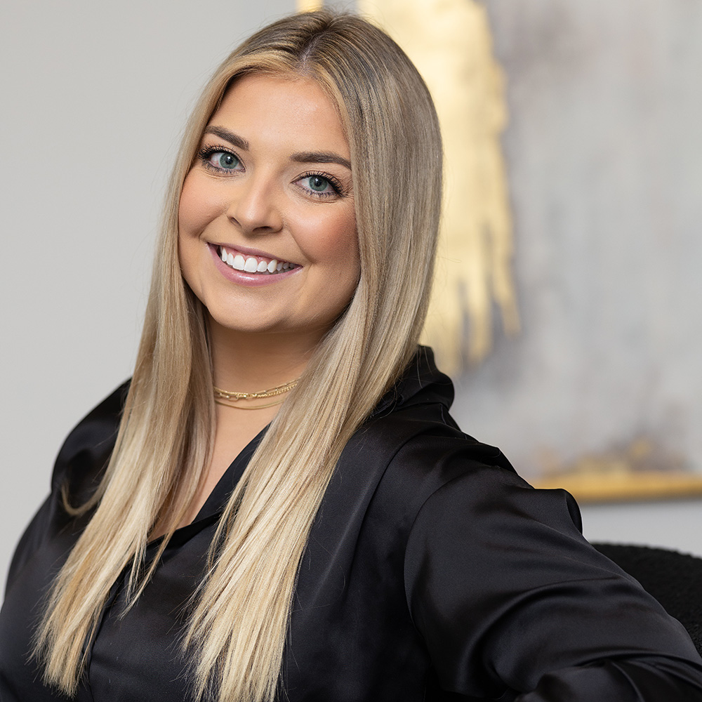 A professional headshot of Lexie Wanninger, a smiling young woman with straight, long blonde hair. She is wearing a silky black button-down shirt and a delicate gold layered necklace. She is looking directly at the camera with a warm smile, positioned against a neutral grey wall featuring a blurred piece of artwork with gold accents.