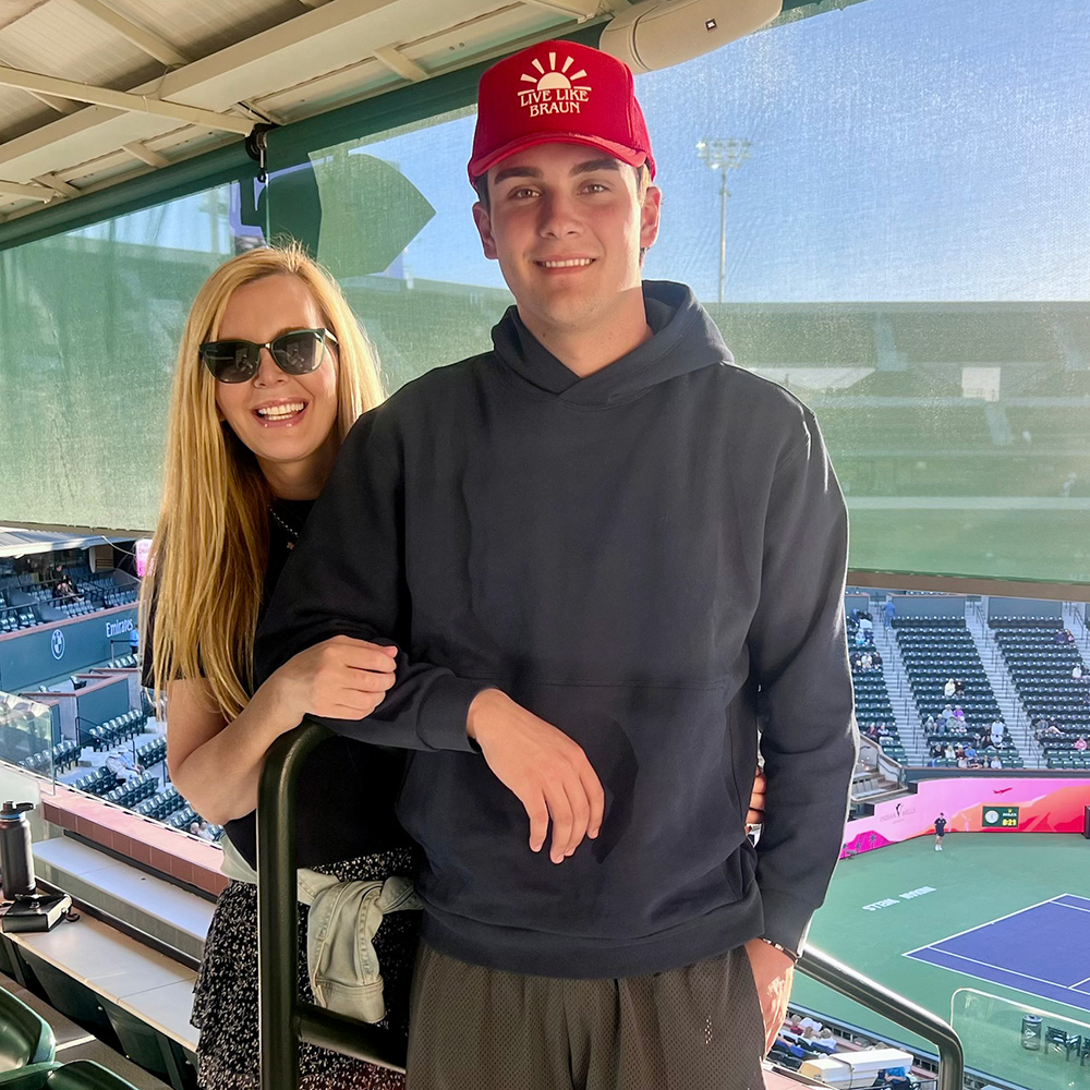 Susan Zinn and her son Jackson Zinn pose for a smiling portrait at the 2026 BNP Paribas Open. Susan, on the left, has long blonde hair and is wearing sunglasses and a black top. She has her hand resting on the arm of Jackson, who is wearing a navy blue hoodie and a red baseball cap with a "Live Like Braun" logo. They are standing in an elevated viewing area, with the vast seating and the purple and green stadium court of the Indian Wells Tennis Garden visible in the background below.