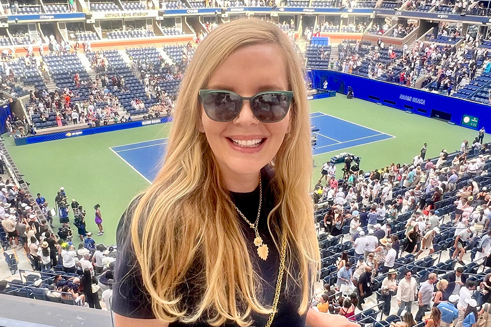 Susan Zinn smiles for a photo from an elevated vantage point at Arthur Ashe Stadium during the US Open. She is wearing dark sunglasses, a black short-sleeved top, and a gold necklace with a leaf-shaped pendant. A gold chain strap from her bag crosses her shoulder. Behind her, the blue and green tennis court is visible, surrounded by thousands of spectators in the stands under the bright afternoon sun. The stadium's upper tiers and digital displays are also visible in the background.