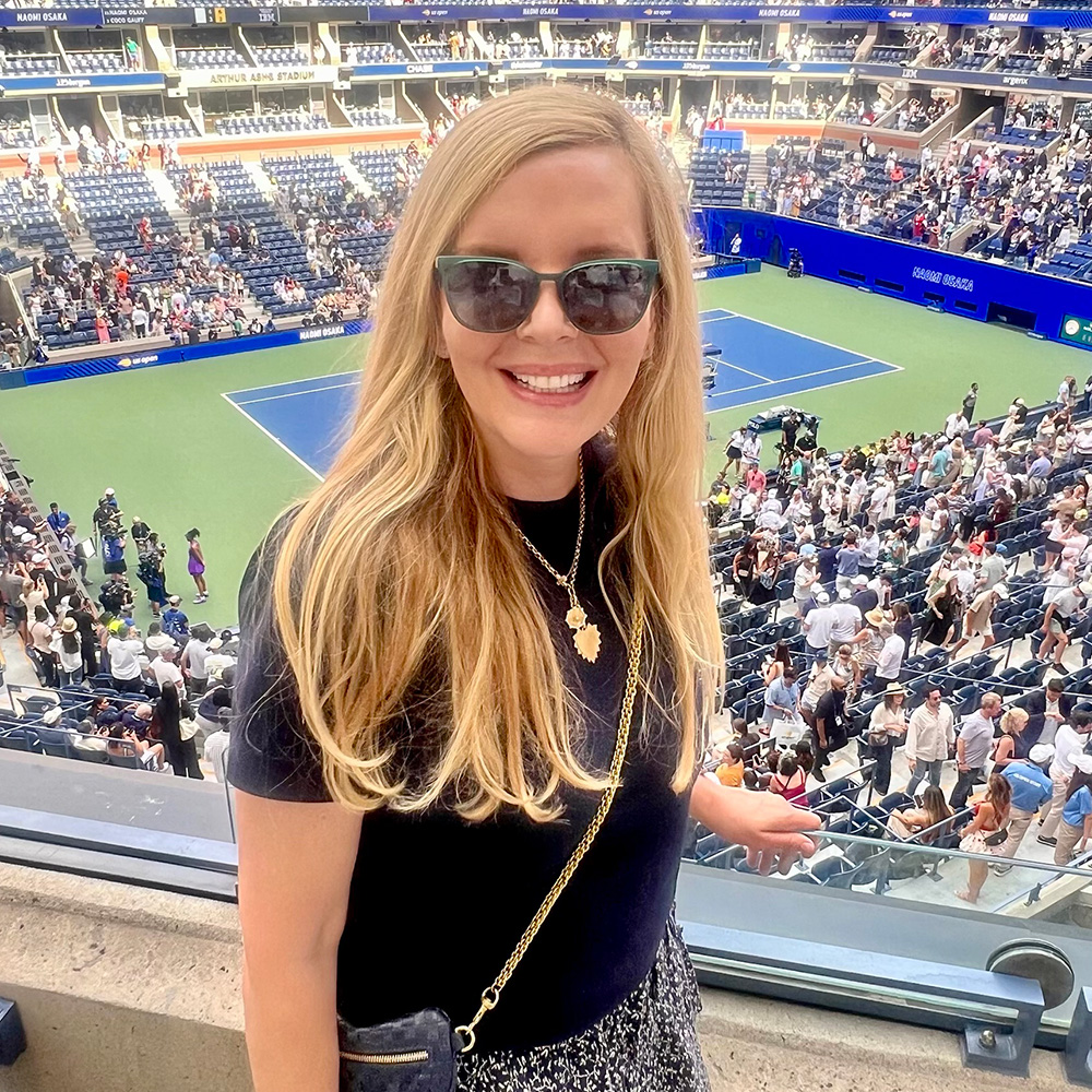Susan Zinn smiles for a photo from an elevated vantage point at Arthur Ashe Stadium during the US Open. She is wearing dark sunglasses, a black short-sleeved top, and a gold necklace with a leaf-shaped pendant. A gold chain strap from her bag crosses her shoulder. Behind her, the blue and green tennis court is visible, surrounded by thousands of spectators in the stands under the bright afternoon sun. The stadium's upper tiers and digital displays are also visible in the background.