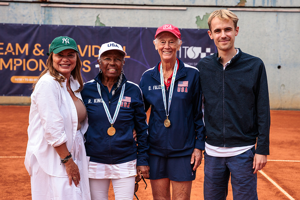 World Doubles champions Roz King and Donna Fales pose on a clay court with two others. Roz (second from left) wears a white "USA" cap and a navy Team USA jacket, while Donna (third from left) wears a red "USA" cap. Both women are wearing their gold medals and smiling for the camera.