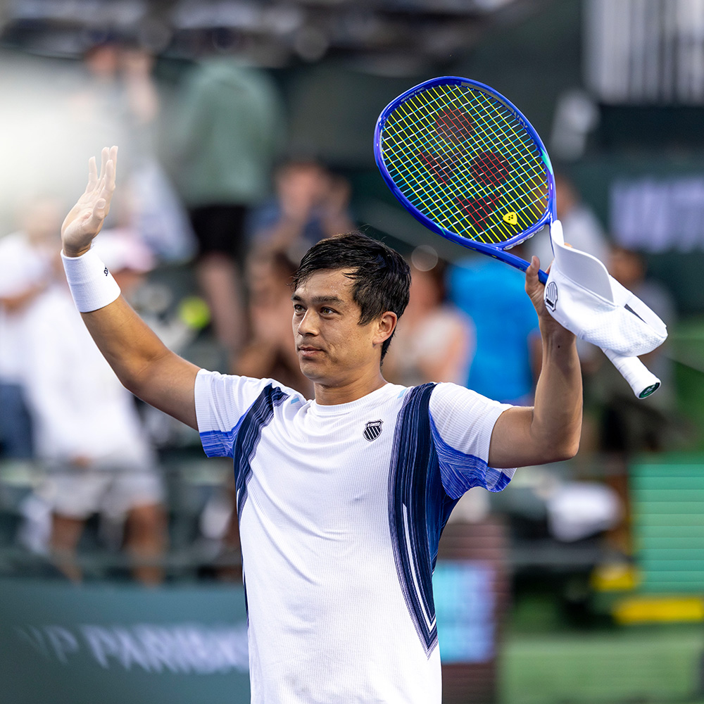 American tennis player Mackenzie McDonald waves to the crowd while celebrating a victory at the 2026 BNP Paribas Open. He is wearing a white and navy blue K-Swiss athletic shirt and a white wristband on his right arm. In his left hand, he triumphantly holds a bright blue Yonex tennis racket and a white towel. The stadium background is filled with blurred spectators, and the BNP Paribas logo is visible on the court barrier behind him.