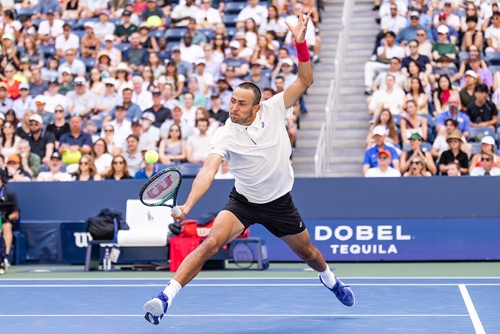American tennis player Emilio Nava lunges across the blue hard court to hit a low backhand during the 2025 US Open. He is shown in full athletic motion, wearing a white Asics polo, black shorts, blue tennis shoes, and a bright pink wristband on his left arm. The background is filled with a crowded stadium of spectators, with a prominent "Dobel Tequila" sign visible on the court-side barrier.