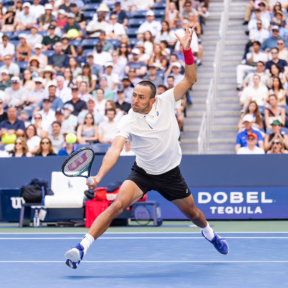 American tennis player Emilio Nava lunges across the blue hard court to hit a low backhand during the 2025 US Open. He is shown in full athletic motion, wearing a white Asics polo, black shorts, blue tennis shoes, and a bright pink wristband on his left arm. The background is filled with a crowded stadium of spectators, with a prominent "Dobel Tequila" sign visible on the court-side barrier.
