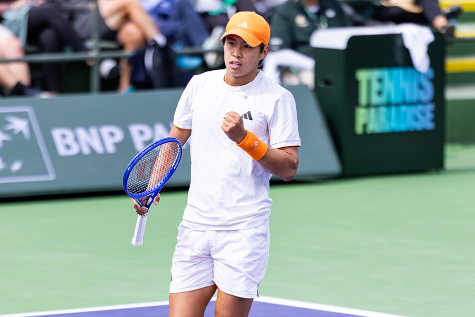 Tennis player Learner Tien celebrates with a fist pump on a purple hard court during the 2026 BNP Paribas Open. He is wearing a white Adidas t-shirt and shorts, paired with a bright orange cap, matching wristbands, and orange socks. He holds a blue tennis racket in his right hand while looking intently ahead. The background shows the green tournament stadium walls featuring the BNP Paribas and "Tennis Paradise" logos, with spectators blurred in the stands.