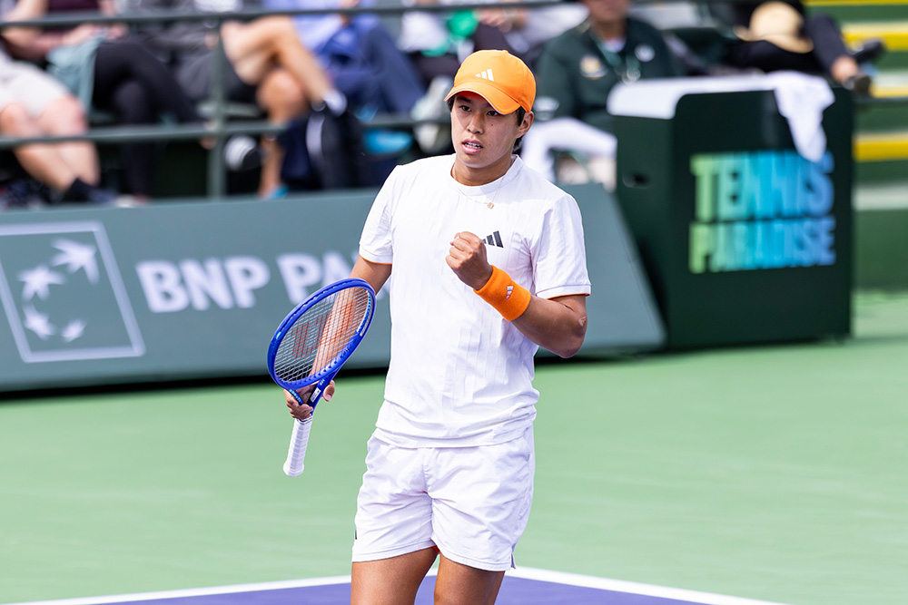 Tennis player Learner Tien celebrates with a fist pump on a purple hard court during the 2026 BNP Paribas Open. He is wearing a white Adidas t-shirt and shorts, paired with a bright orange cap, matching wristbands, and orange socks. He holds a blue tennis racket in his right hand while looking intently ahead. The background shows the green tournament stadium walls featuring the BNP Paribas and "Tennis Paradise" logos, with spectators blurred in the stands.