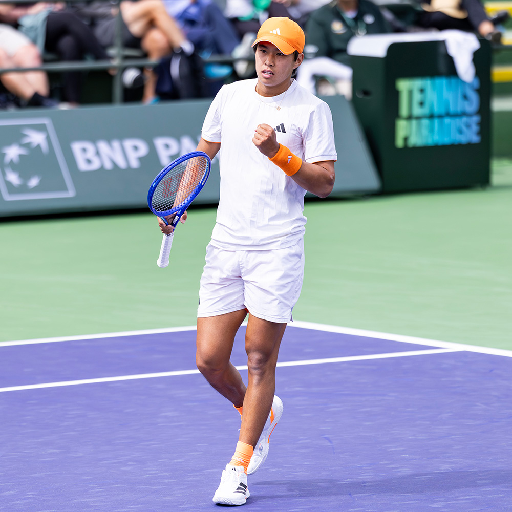 Tennis player Learner Tien celebrates with a fist pump on a purple hard court during the 2026 BNP Paribas Open. He is wearing a white Adidas t-shirt and shorts, paired with a bright orange cap, matching wristbands, and orange socks. He holds a blue tennis racket in his right hand while looking intently ahead. The background shows the green tournament stadium walls featuring the BNP Paribas and "Tennis Paradise" logos, with spectators blurred in the stands.