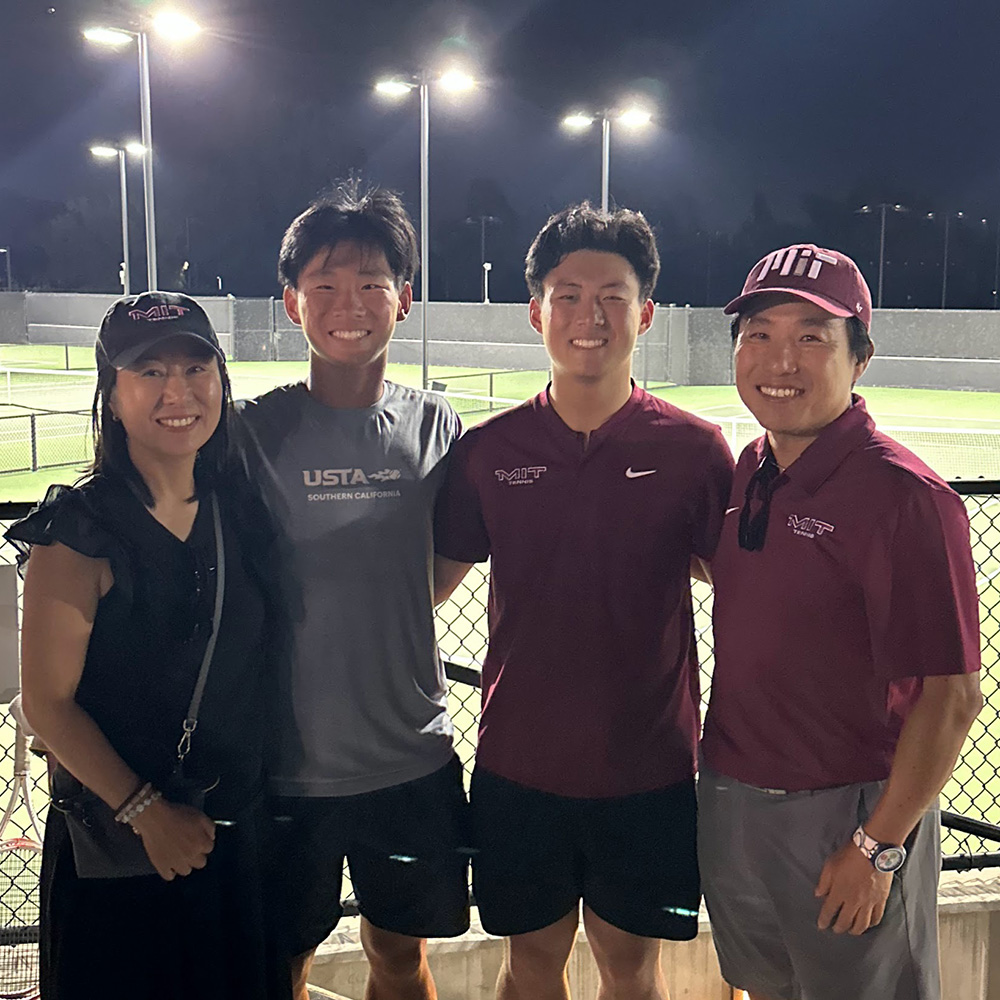 A group portrait of Dr. KyoungBin Im and his family smiling together at night on a tennis court in March 2026. From left to right: a woman in a black top and an MIT Tennis cap, a young man in a grey USTA Southern California t-shirt, a young man in a maroon MIT Tennis shirt, and Dr. Im wearing a maroon MIT Tennis polo and matching cap. Bright stadium lights illuminate the courts in the background behind a chain-link fence.