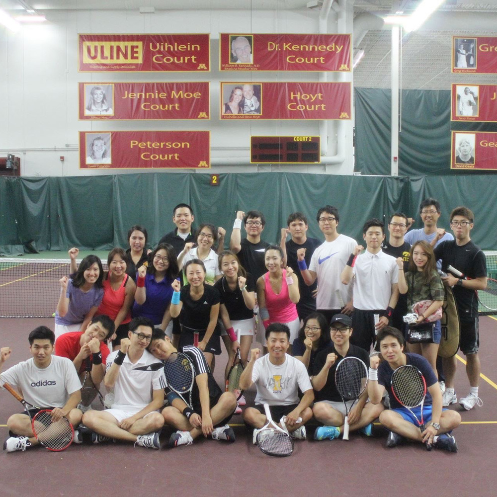 A large group portrait of Dr. KyoungBin Im and his college tennis club team posing on a court in Chicago, September 2016. The team members are arranged in rows, with several players sitting in the front holding rackets and others standing behind them, many raising their fists in a celebratory gesture. Dr. Im is seated in the front row toward the center, wearing a white t-shirt and smiling. In the background, maroon banners hang on the wall identifying various courts, such as Uihlein Court and Jennie Moe Court.