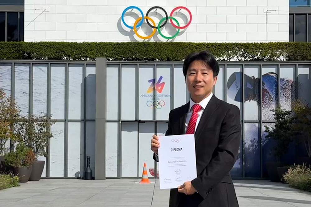 Dr. KyoungBin Im stands proudly in Lausanne, Switzerland, holding his IOC Diploma in Sports Psychiatry in September 2025. He is dressed in a black suit with a bright red tie, posing in an open plaza. Behind him is a modern building featuring the iconic Olympic rings and a "Milano Cortina 2026" logo on the glass doors. The scene is bright and sunny, with his shadow cast on the grey stone pavement.