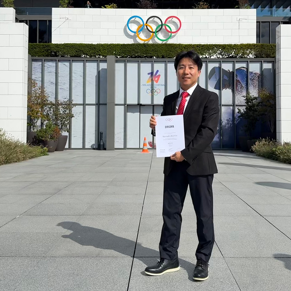 Dr. KyoungBin Im stands proudly in Lausanne, Switzerland, holding his IOC Diploma in Sports Psychiatry in September 2025. He is dressed in a black suit with a bright red tie, posing in an open plaza. Behind him is a modern building featuring the iconic Olympic rings and a "Milano Cortina 2026" logo on the glass doors. The scene is bright and sunny, with his shadow cast on the grey stone pavement.