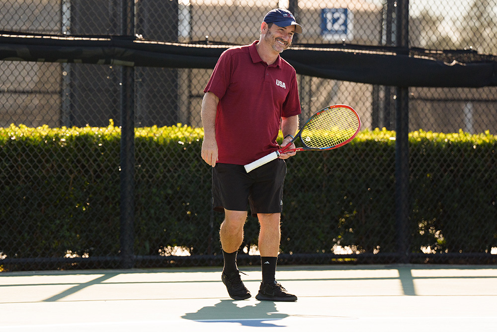 Jason Harnett instructs wheelchair tennis players during a clinic at the USTA National Campus in Orlando, standing courtside with players gathered around him on an outdoor tennis court.