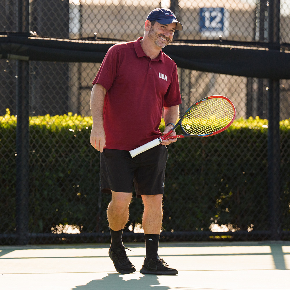 Jason Harnett instructs wheelchair tennis players during a clinic at the USTA National Campus in Orlando, standing courtside with players gathered around him on an outdoor tennis court.