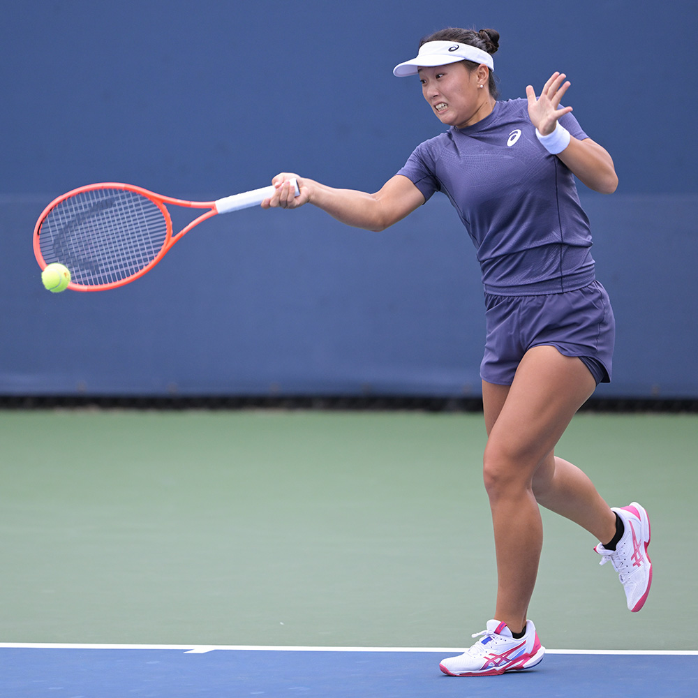 Tennis player Claire Liu hits a powerful forehand during a women's qualifying singles match at the 2025 US Open. She is captured in mid-motion on a green and blue hard court, wearing a charcoal grey Asics athletic top and matching shorts, a white visor, and white tennis shoes with pink accents. She holds an orange racket, focused intently on the ball. The background features a solid blue stadium wall.