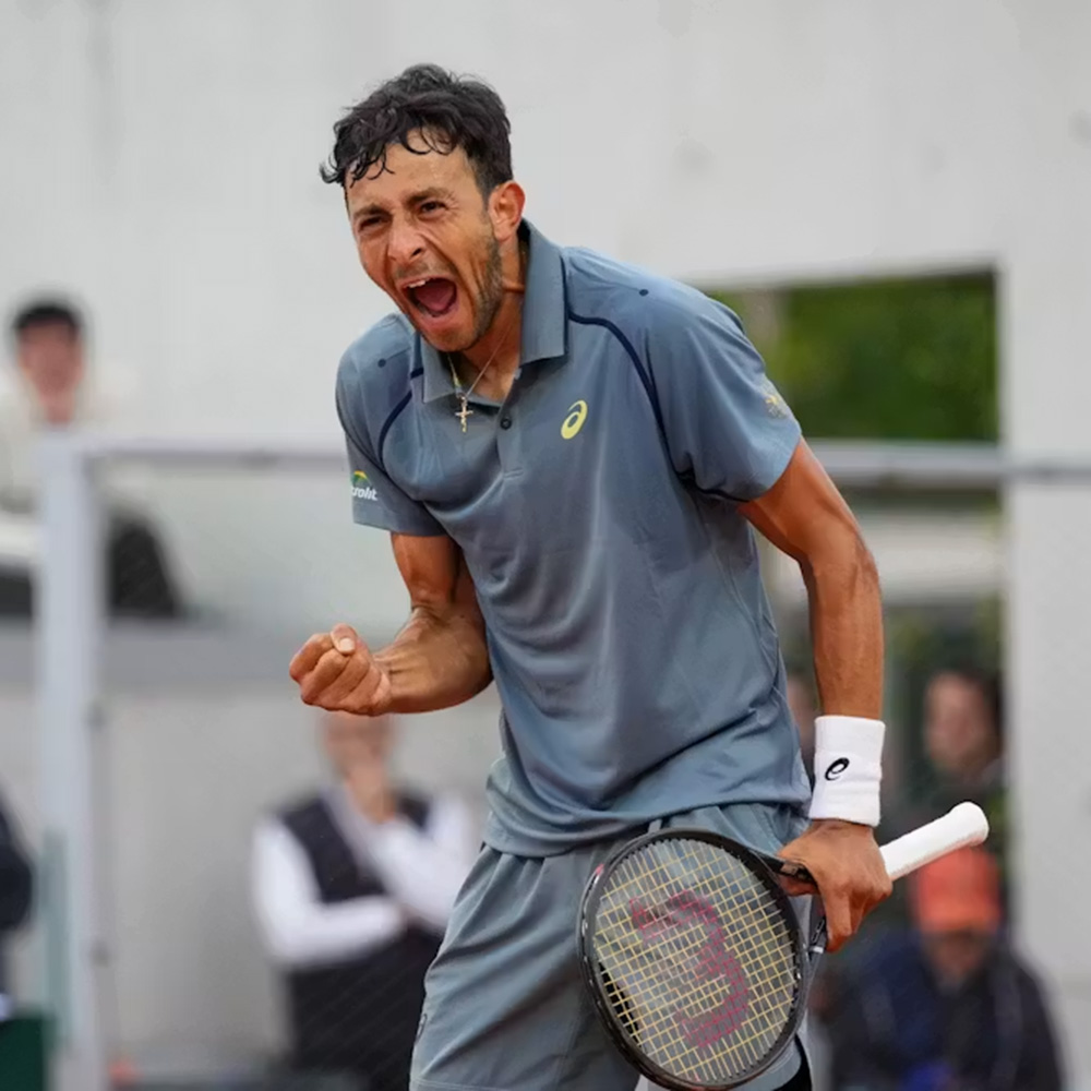 Tennis player Emilio Nava lets out a powerful shout and pumps his fist in celebration during a qualifying match at Roland Garros. He is wearing a grey Asics polo shirt with navy trim and a white wristband on his left arm. He holds his tennis racket in his left hand, appearing intense and focused after a big point. The background shows the blurred figures of spectators and officials at the stadium.