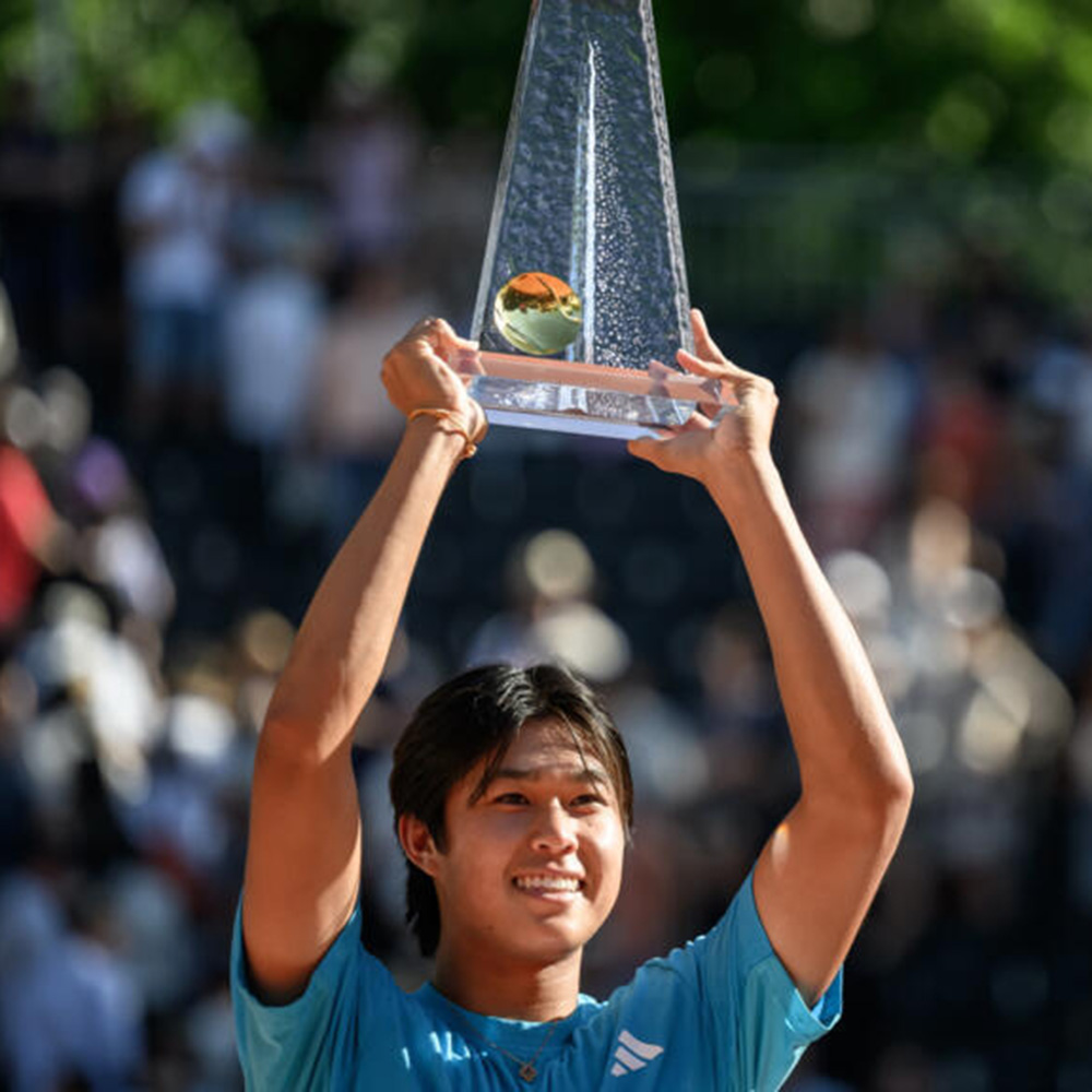 Tennis player Learner Tien smiles broadly while holding the Geneva Open glass trophy high above his head in celebration. He is wearing a turquoise Adidas athletic shirt. The background is filled with the blurred figures of a cheering crowd in stadium seating.