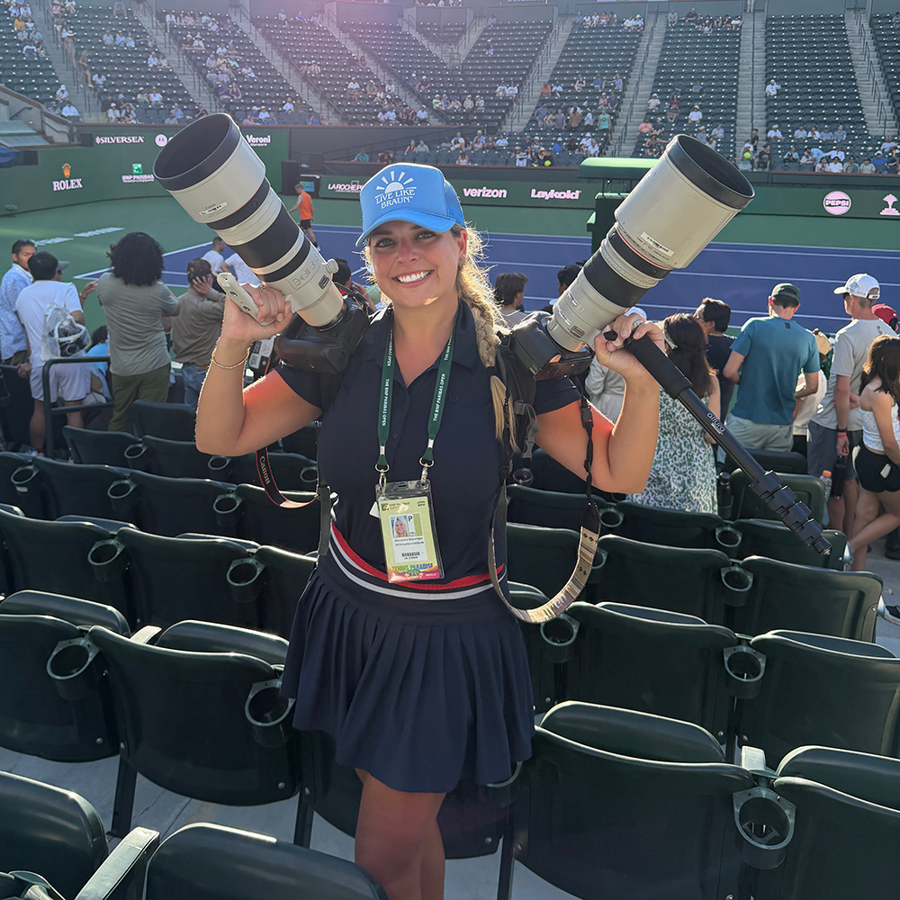 A portrait of photographer Lexie Wanninger smiling broadly while working at the BNP Paribas Open. She is standing in the stadium stands, wearing a navy blue polo and matching pleated skirt, a bright blue "Live Like Braun" trucker hat, and a tournament media credential around her neck. She is playfully balancing two large professional DSLR cameras with massive white telephoto lenses on her shoulders. The background shows the blue and green tennis stadium court with blurred spectators and sponsor banners like Rolex and Verizon.