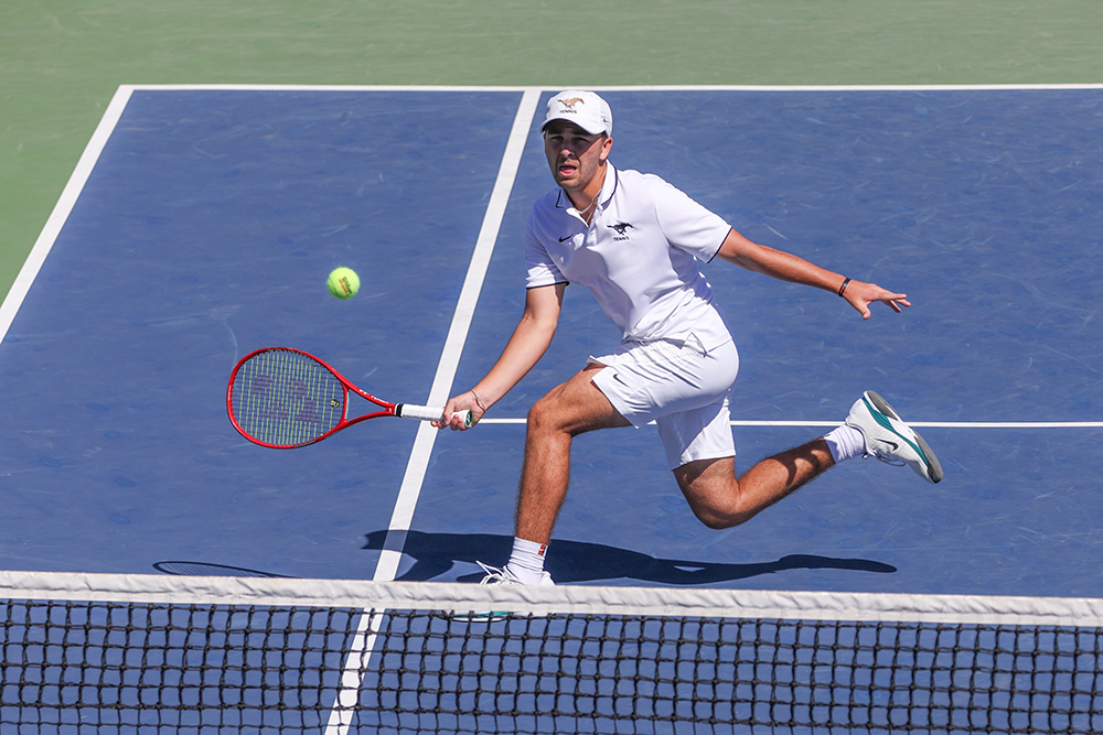 Trevor Svajda lunges across a blue outdoor hard court to hit a low forehand. He is wearing an all-white SMU Tennis uniform, including a polo shirt, shorts, and cap. The action shot is taken from an elevated angle behind the net, showing him in full stride as he reaches for the ball with his red racket.
