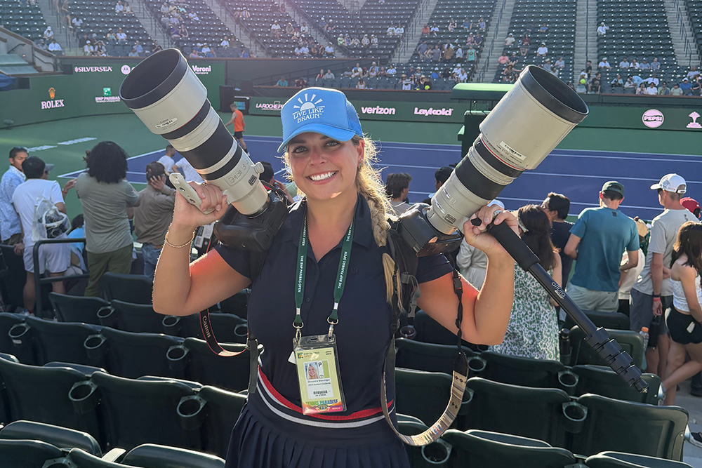 A portrait of photographer Lexie Wanninger smiling broadly while working at the BNP Paribas Open. She is standing in the stadium stands, wearing a navy blue polo and matching pleated skirt, a bright blue "Live Like Braun" trucker hat, and a tournament media credential around her neck. She is playfully balancing two large professional DSLR cameras with massive white telephoto lenses on her shoulders. The background shows the blue and green tennis stadium court with blurred spectators and sponsor banners like Rolex and Verizon.