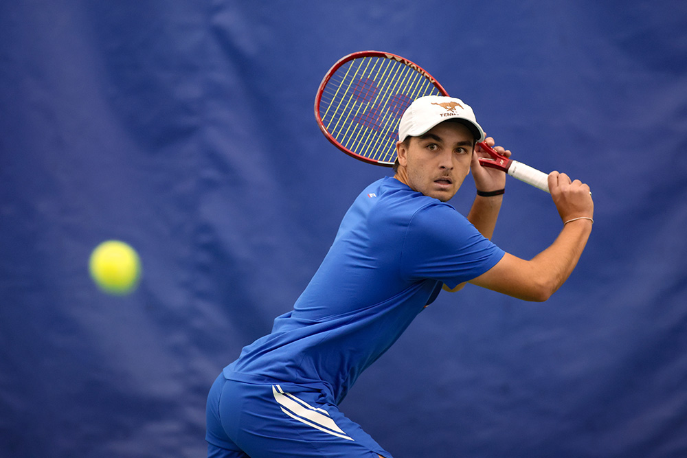 Tennis player Trevor Svajda prepares for a two-handed backhand during an indoor match. He is wearing a blue athletic shirt and a white SMU Tennis cap, tracking a yellow tennis ball in mid-air. He holds a red Yonex racket against a solid blue backdrop.