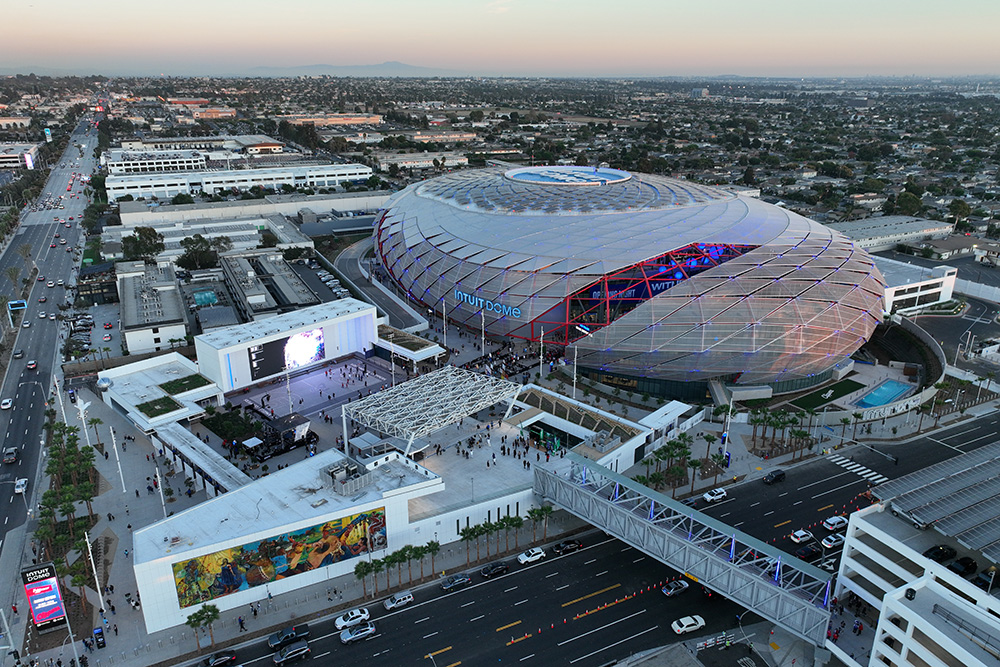An aerial, high-angle shot of the Intuit Dome arena and its surrounding campus during dusk. The main arena features a distinct, domed geometric shell structure illuminated with subtle blue and red accent lights and displays the "INTUIT DOME" sign on its exterior wall. An outdoor plaza, complete with a large video board, an open courtyard, a pedestrian skybridge crossing a busy multi-lane street, and palm trees, sits adjacent to the arena. In the background, a sprawling cityscape extends to the horizon under a pale evening sky.