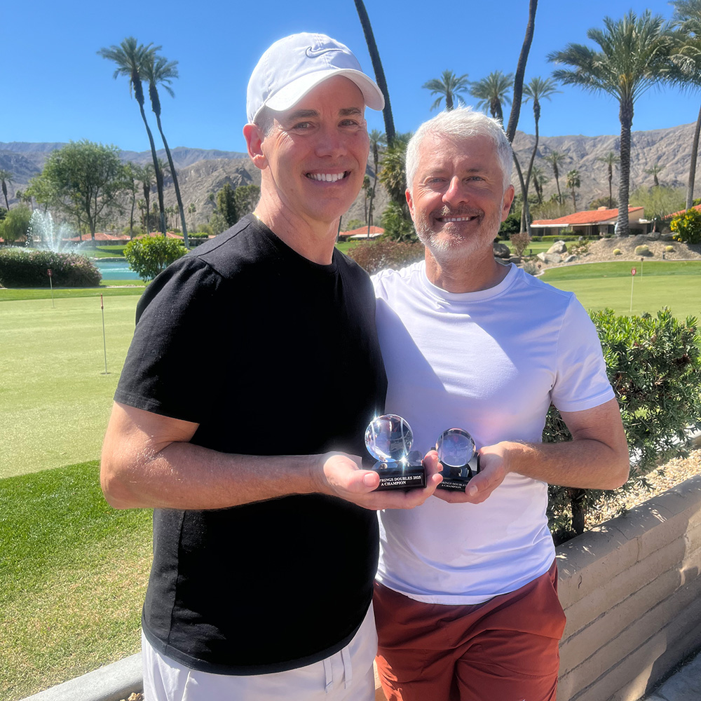 Two smiling men pose together outdoors on a sunny day, each holding a small crystal ball trophy. The man on the left wears a black t-shirt, white shorts, and a white Nike cap. The man on the right wears a white crew-neck shirt and rust-colored shorts. The scenic background features a lush green golf course, a water fountain, tall palm trees, and desert mountains under a clear blue sky.