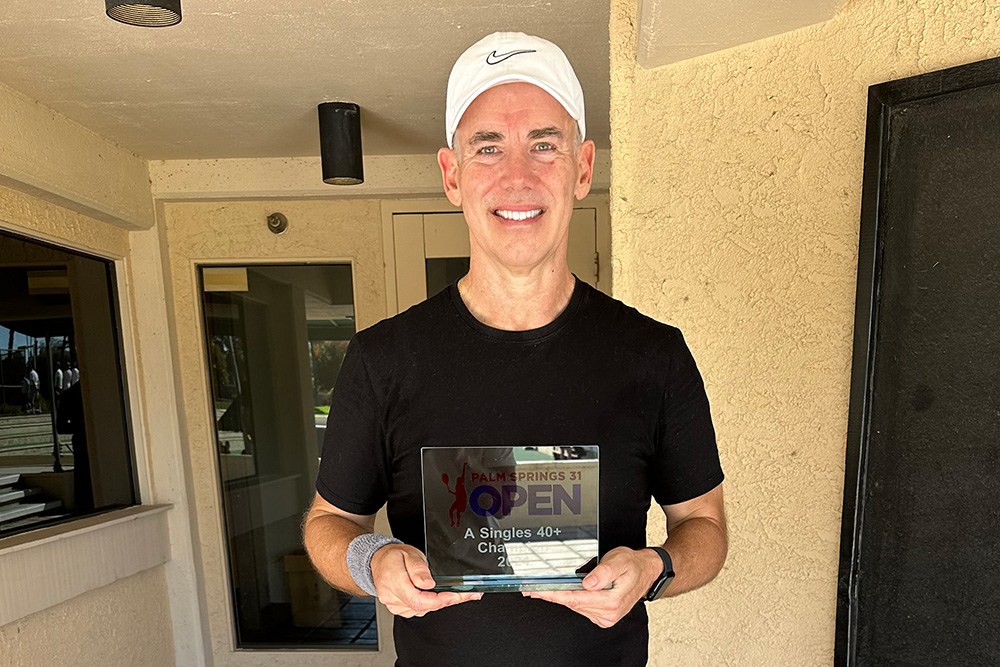 Ralph Nunley holding a glass trophy under a covered entryway. He is wearing a black crew-neck t-shirt, a white Nike baseball cap, a grey wristband on his right arm, and a black smartwatch on his left wrist. He holds the square plaque-style trophy with both hands, which reads "Palm Springs 31 Open - A Singles 40+ Champion 2024." The background consists of a textured, light beige wall and glass office doors.
