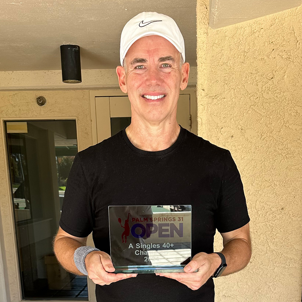 Ralph Nunley holding a glass trophy under a covered entryway. He is wearing a black crew-neck t-shirt, a white Nike baseball cap, a grey wristband on his right arm, and a black smartwatch on his left wrist. He holds the square plaque-style trophy with both hands, which reads "Palm Springs 31 Open - A Singles 40+ Champion 2024." The background consists of a textured, light beige wall and glass office doors.