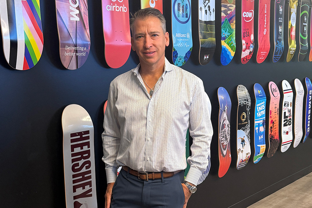 John Lansville poses for a full-length portrait in front of a striking feature wall decorated with custom skateboards. He is smiling slightly with one hand in his pocket, wearing a light-colored patterned dress shirt, grey trousers with a brown leather belt, and brown leather dress shoes. The black wall behind him displays two long rows of skateboard decks, each sporting unique colorful graphic designs and corporate or event branding, including prominent logos for LA28, AECOM, airbnb, and HERSHEY.