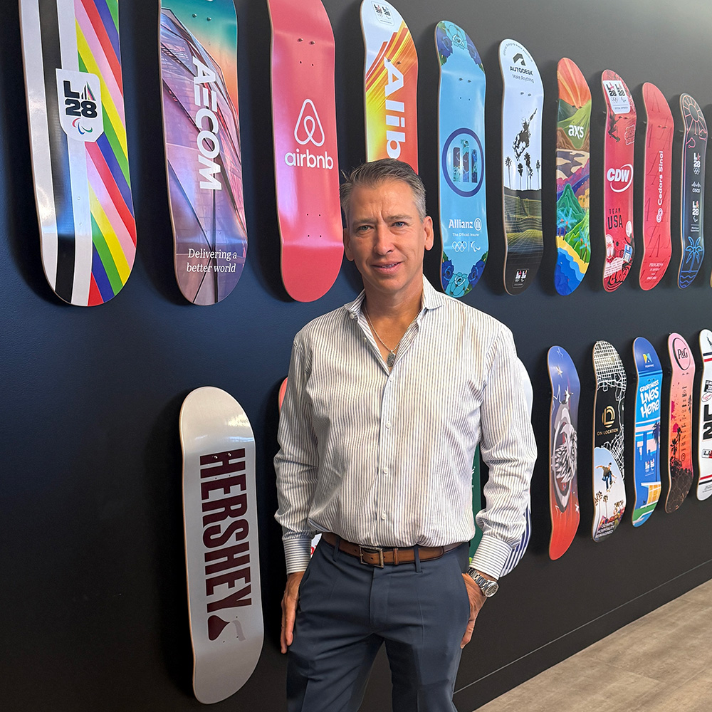 John Lansville poses for a full-length portrait in front of a striking feature wall decorated with custom skateboards. He is smiling slightly with one hand in his pocket, wearing a light-colored patterned dress shirt, grey trousers with a brown leather belt, and brown leather dress shoes. The black wall behind him displays two long rows of skateboard decks, each sporting unique colorful graphic designs and corporate or event branding, including prominent logos for LA28, AECOM, airbnb, and HERSHEY.