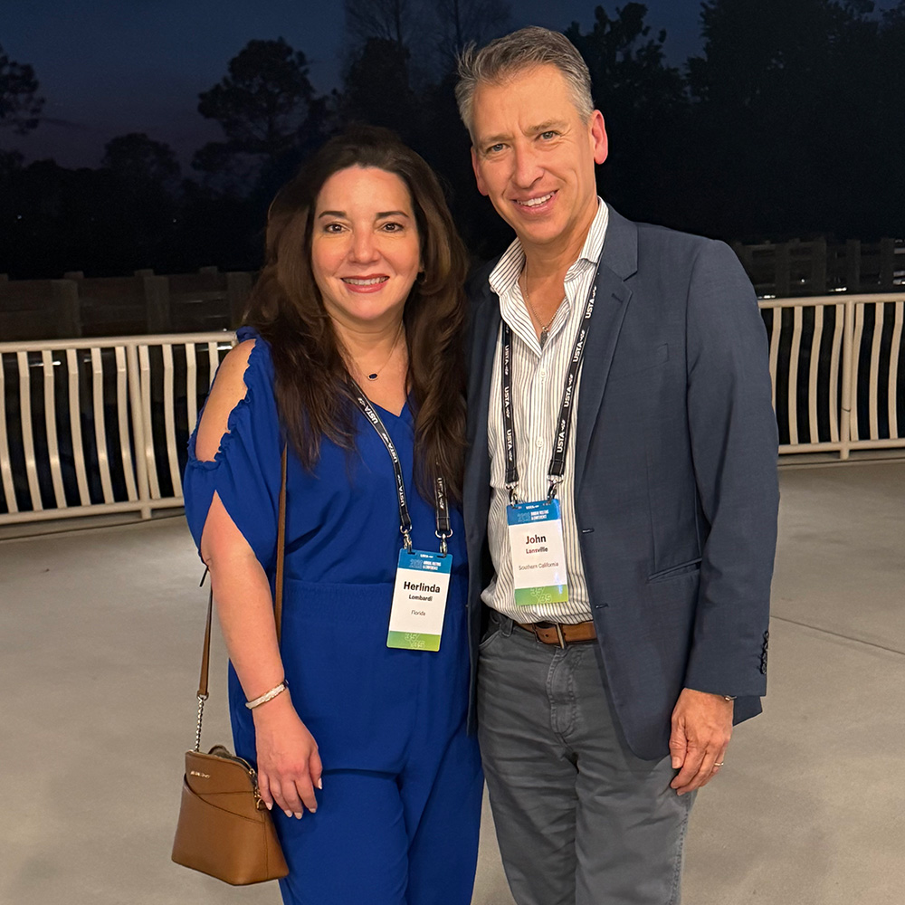Herlinda Lombardi and John Lansville pose together for a photo at the USTA Annual Meeting & Conference. They stand shoulder-to-shoulder outdoors at dusk on a paved balcony or patio with a white railing behind them. Herlinda, on the left, smiles while wearing a royal blue jumpsuit with cold-shoulder cutouts and carrying a brown crossbody purse. John, on the right, smiles alongside her, dressed in a navy blue blazer, a striped button-down shirt, and grey trousers. Both wear official event lanyards around their necks displaying their credentials, which identify Herlinda with USTA Florida and John with USTA Southern California.
