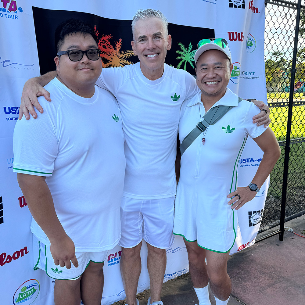 Three men smile for a group photo in front of a white tournament banner. All three are dressed in matching white athletic gear with green trim and a green Adidas-style leaf logo. The man in the center has his arms around the shoulders of the other two men. The man on the right also wears a white visor with green sunglasses resting on top. The background includes a tennis court fence under bright daylight.