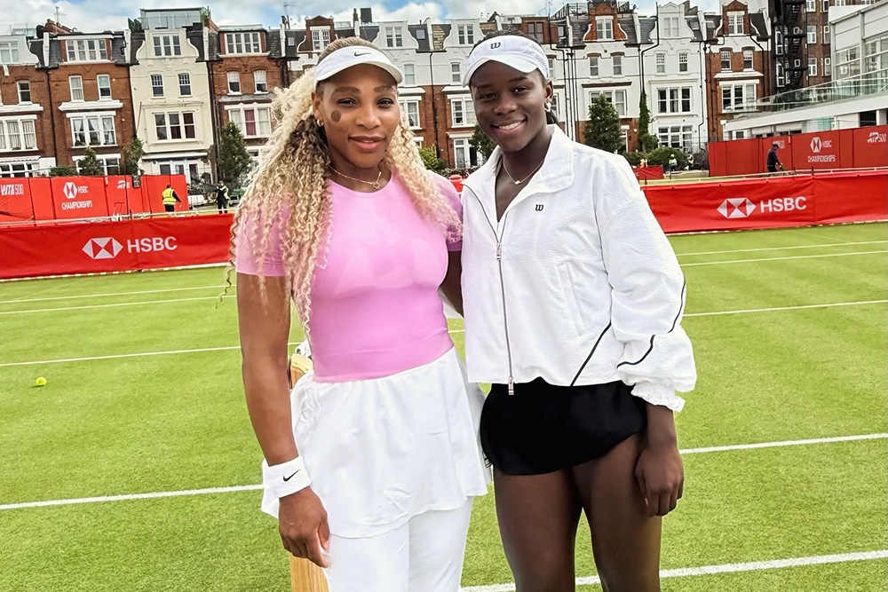 Tennis legend Serena Williams and player Victoria Mboko pose together for a photo on an outdoor grass tennis court. Serena, on the left, smiles while wearing a pink short-sleeved top, a white tennis skirt over white athletic pants, white Nike shoes, and a white visor. Victoria Mboko, on the right, smiles alongside her, dressed in a white Wilson zip-up jacket, black athletic shorts, white tennis shoes, and a white visor. They are standing on the green grass court under a bright, partly cloudy sky, with red HSBC sponsor banners and row houses visible in the background.